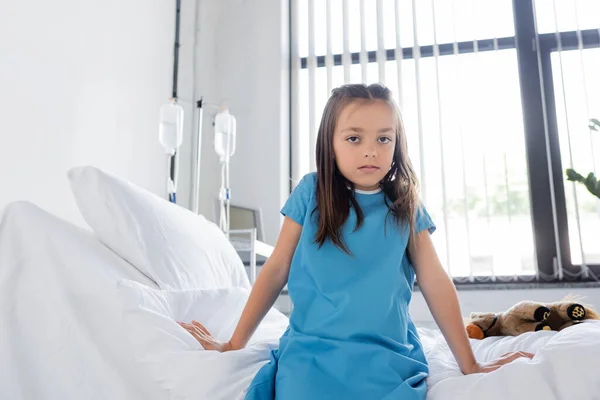 Niño en bata de paciente mirando a la cámara mientras está sentado en la cama en el hospital - foto de stock