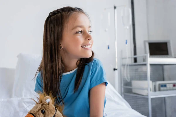 Позитивный ребенок в больничном халате смотрит в сторону мягкой игрушки на кровати в клинике — стоковое фото