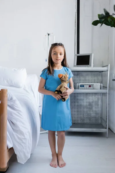 Enfant pieds nus en robe de patient tenant un jouet doux près du lit dans la salle d'hôpital — Photo de stock