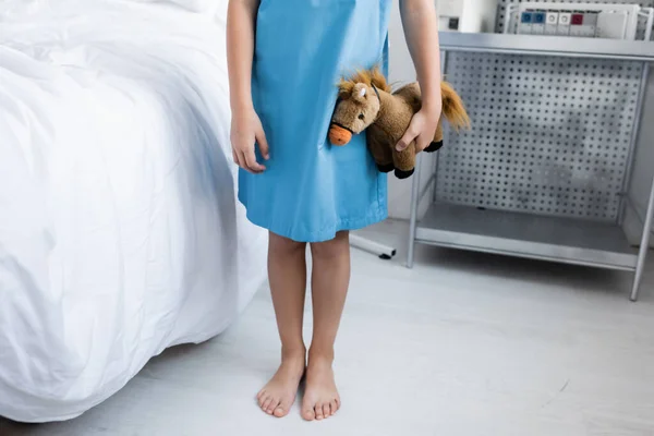 Cropped view of barefoot child in patient gown holding soft toy in hospital ward — Stock Photo