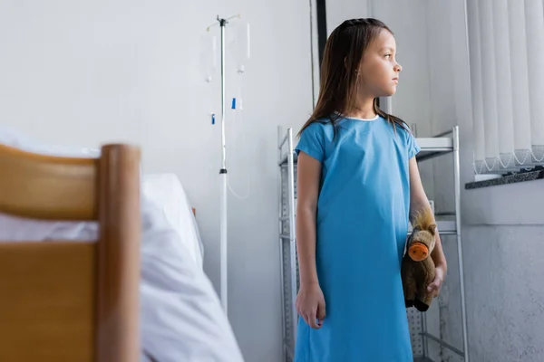 Вид сбоку ребенка в халате, держащего мягкую игрушку рядом с кроватью в больничной палате — стоковое фото