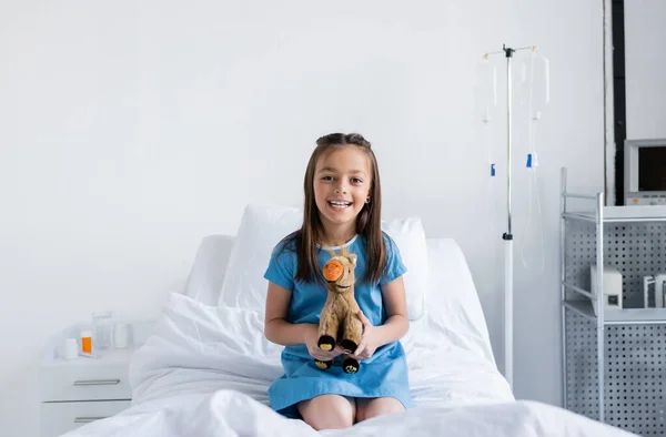 Улыбающаяся девушка в больничном халате держит мягкую игрушку и смотрит в камеру в больничном отделении — стоковое фото