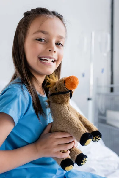 Retrato de menina sorridente em vestido de paciente segurando brinquedo na enfermaria hospital borrada — Fotografia de Stock
