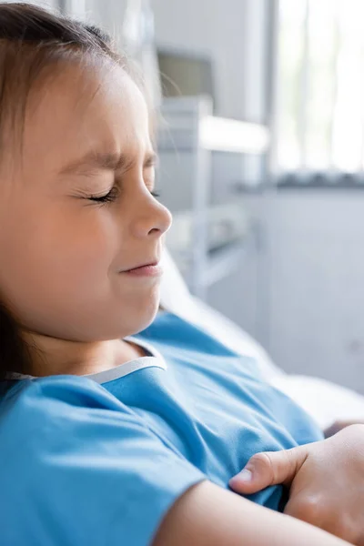 Niño disgustado y enfermo en bata de paciente acostado en la cama en la clínica - foto de stock