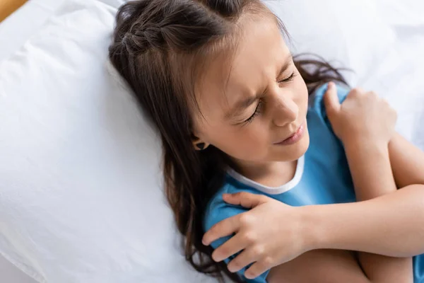 Вид сверху на больного ребенка, трогающего плечи на кровати в клинике — стоковое фото