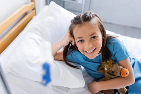 Vista de ángulo alto de niña alegre sosteniendo juguete en la cama en la clínica - foto de stock