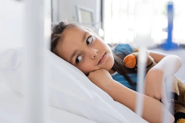 Niño abrazando juguete suave mientras está acostado en la cama en la sala de hospital - foto de stock