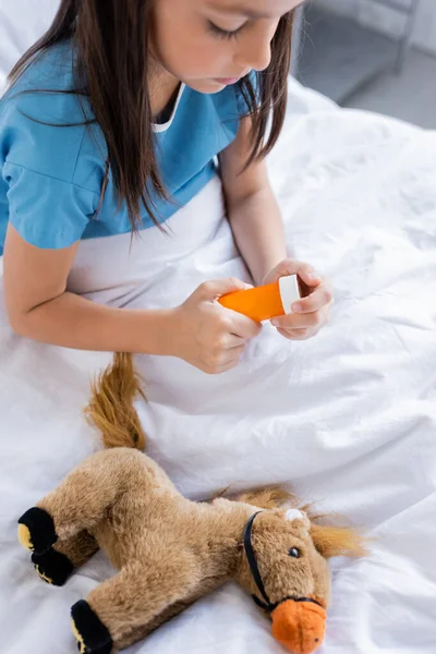 Criança em vestido de paciente segurando garrafa com pílulas perto de brinquedo macio na cama do hospital — Fotografia de Stock