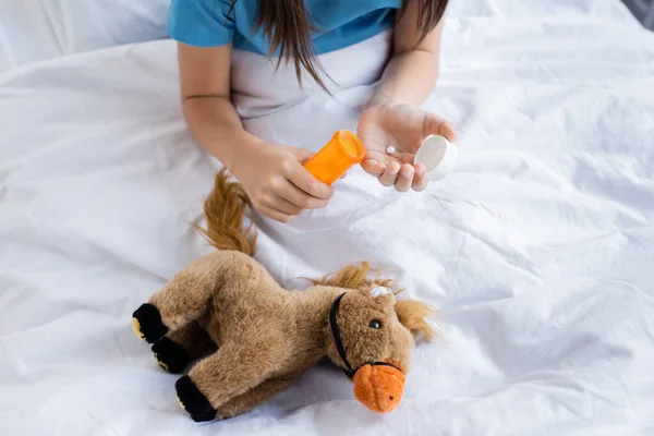 Обрізаний вид на дитину, що тримає таблетки біля іграшки на ліжку в клініці — стокове фото