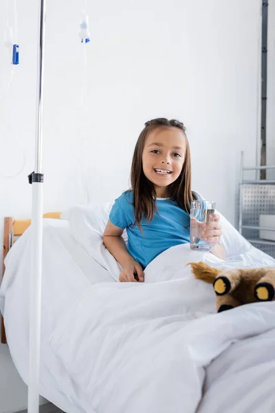 Chica sonriente en bata de paciente sosteniendo un vaso de agua en la cama en el hospital - foto de stock