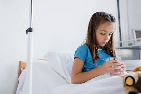Ill criança segurando vidro de água perto de brinquedo borrado na cama na clínica — Fotografia de Stock