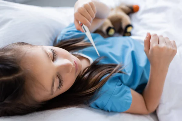 Mädchen im Patientenkleid hält verschwommenes elektronisches Thermometer auf Bett in Klinik — Stockfoto