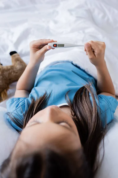 Criança segurando termômetro eletrônico enquanto estava deitada na cama na enfermaria do hospital — Fotografia de Stock