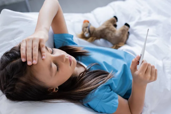 Sick kid holding electronic thermometer and touching forehead on bed in clinic — Stock Photo