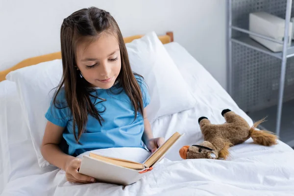 Child reading book near soft toy on hospital bed — Stock Photo