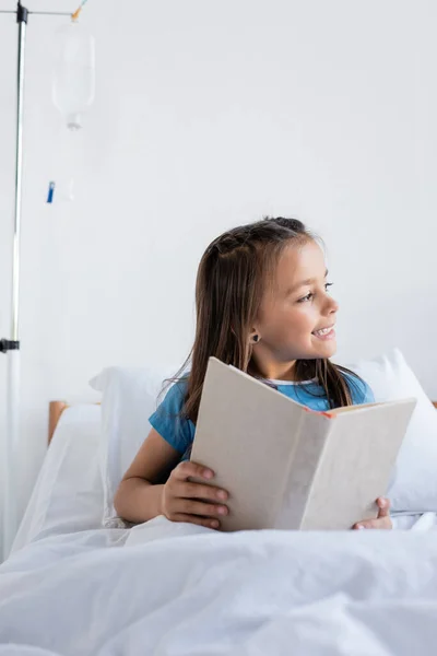 Criança sorridente segurando livro na cama na enfermaria do hospital — Fotografia de Stock