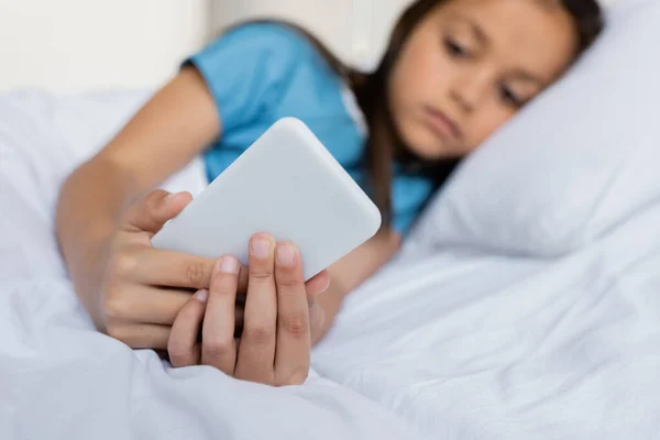 Niño borroso usando el teléfono celular mientras está acostado en la cama del hospital - foto de stock