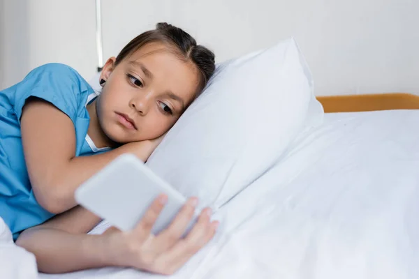 Kid in patient gown using blurred smartphone on hospital bed — Stock Photo