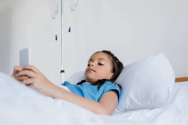 Niño enfermo usando teléfono móvil cerca de terapia intravenosa borrosa en el hospital - foto de stock