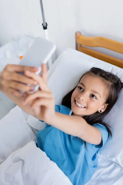 Vista de ángulo alto del niño sonriente tomando selfie en el teléfono inteligente en la sala de hospital - foto de stock
