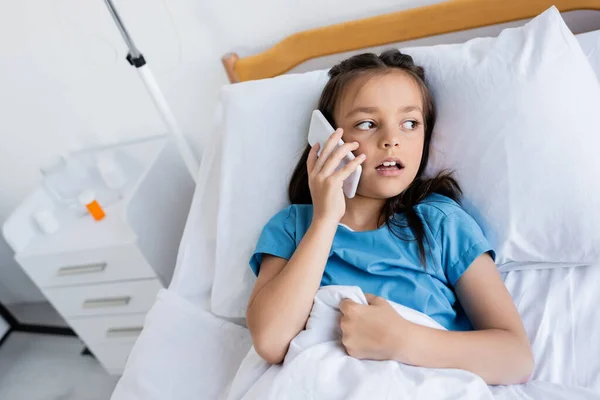 Niño enfermo hablando en el teléfono inteligente mientras está acostado en la cama en la clínica - foto de stock