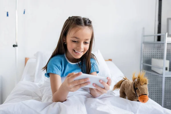 Chica sonriente usando teléfono inteligente cerca de juguete suave en la cama en la clínica - foto de stock