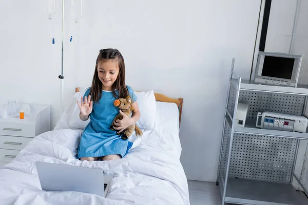 Улыбающаяся девушка держит мягкую игрушку и машет рукой во время видеозвонка на ноутбук в больнице — стоковое фото