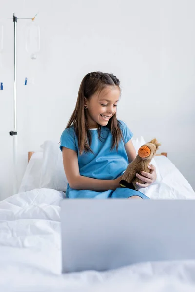 Bambino allegro che tiene il giocattolo vicino al computer portatile offuscato sul letto d'ospedale — Foto stock