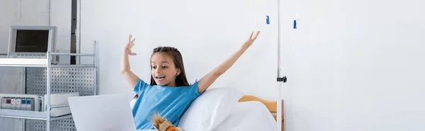 Niño emocionado en bata de paciente mirando a la computadora portátil en la sala de hospital, pancarta - foto de stock