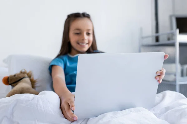Blurred kid in patient gown using laptop near toy on bed in hospital — Stock Photo