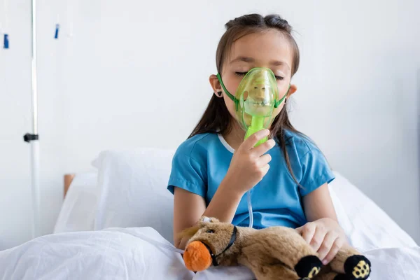 Больной ребенок использует кислородную маску рядом с игрушкой на кровати в больнице — стоковое фото