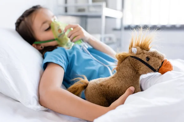 Blurred girl with oxygen mask touching soft toy on bed in clinic — Stock Photo