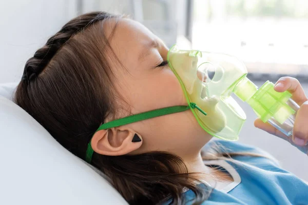 Diseased kid holding oxygen mask while lying on bed in clinic — Stock Photo