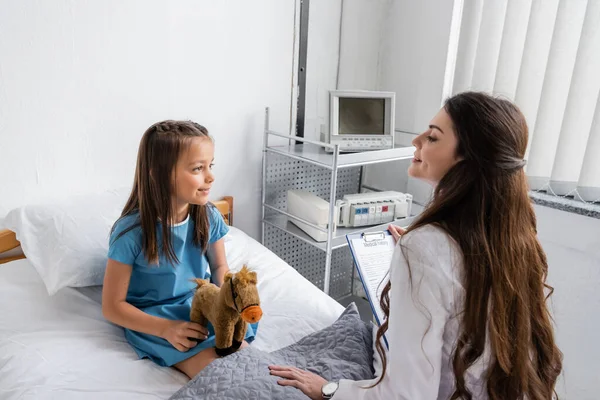 Smiling doctor holding clipboard and talking to child with toy on bed in hospital — Stock Photo
