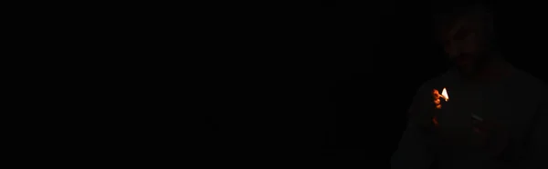 Homem com caixa de fósforos olhando para chama de fósforo iluminado isolado em preto, banner — Fotografia de Stock