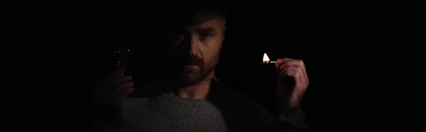 Людина з лампочкою і запаленим сірником дивиться на камеру під час відключення енергії ізольовано на чорному, банер — стокове фото