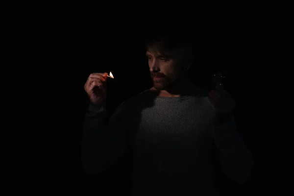 Man in darkness holding electric bulb and looking at flame of lit match isolated on black — Stock Photo
