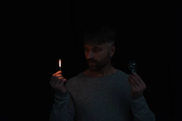 Hombre mirando la llama del fósforo encendido mientras sostiene la bombilla eléctrica aislada en negro - foto de stock