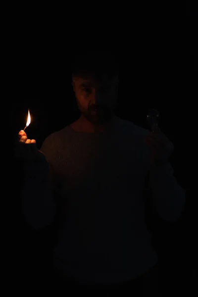 Man with electric bulb and burning match looking at camera in darkness isolated on black — Stock Photo