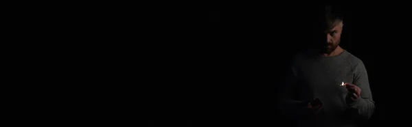 Незадоволений чоловік з сірниковою коробкою і палаючим сірником, дивлячись на камеру під час відключення електроенергії ізольовано на чорному, банер — стокове фото