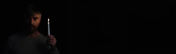 Homem segurando vela acesa e olhando para a câmera durante o apagão de eletricidade isolado em preto, banner — Fotografia de Stock