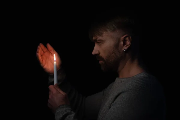 Seitenansicht eines Mannes, der die Hand in der Nähe der Flamme einer brennenden Kerze hält, isoliert auf schwarz — Stockfoto