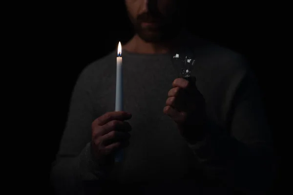 Cropped view of man holding light bulb and lit candle during energy blackout isolated on black — Stock Photo