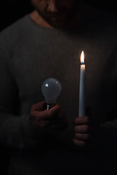 Cropped view of man in darkness holding lit candle and electric bulb isolated on black — Stock Photo