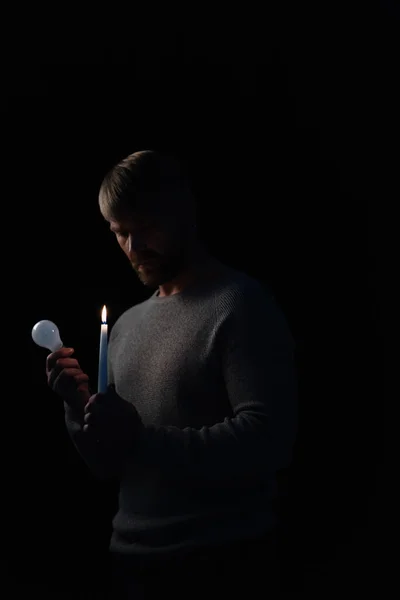 Adult man holding bulb and lit candle during electricity shutdown isolated on black — Stock Photo