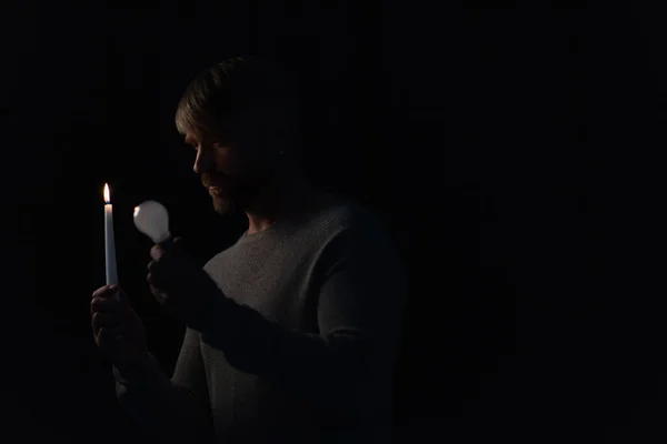 Homme tenant ampoule et bougie allumée dans l'obscurité isolé sur noir — Photo de stock