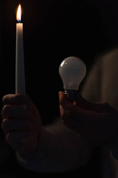 Partial view of man with light bulb and lit candle during power outage isolated on black — Stock Photo