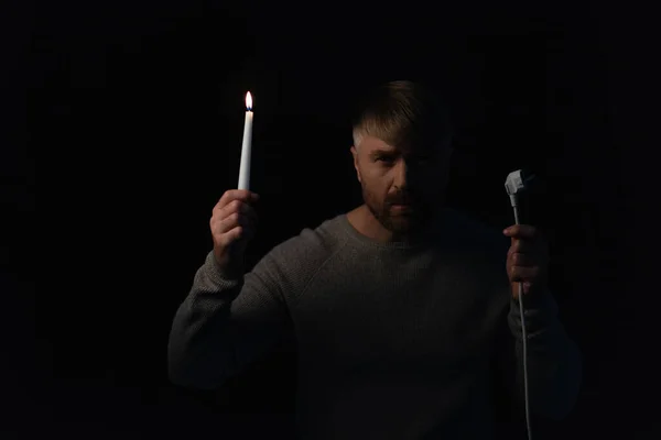 Mann mit Stecker und brennender Kerze schaut während Stromabschaltung isoliert auf schwarz in die Kamera — Stockfoto