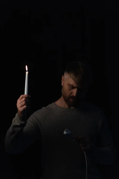 Человек с зажженной свечой, смотрящий на электрическую розетку во время отключения энергии изолирован на черном — стоковое фото