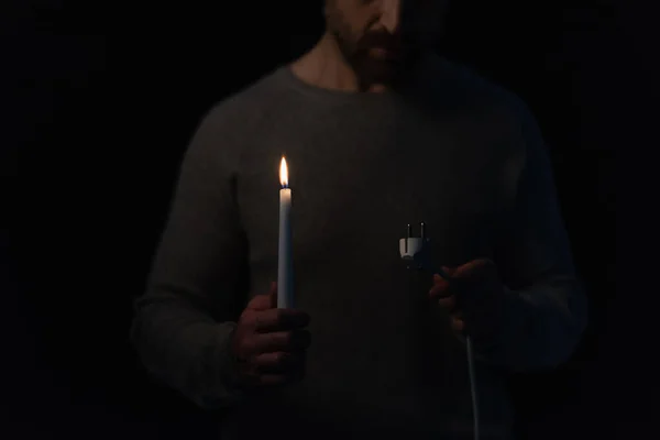 Partial view of man looking at electric plug while holding burning candle during power blackout isolated on black — Stock Photo